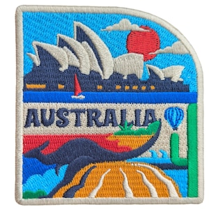 Op de afbeelding: Geborduurde patch met het woord "AUSTRALIA" en Australische symbolen. Het ontwerp omvat het Sydney Opera House, een kangoeroe, een luchtballon en een rode zon tegen een blauwe lucht. De patch heeft een witte rand.