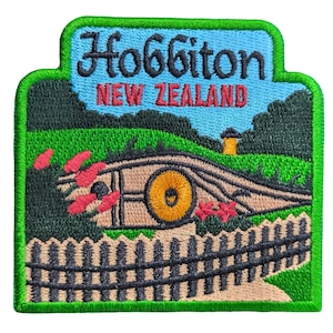 以下が含まれることがあります： 青い背景に「Hobbiton」と「New Zealand」の文字が刺繍されたパッチ。丸い窓、フェンス、緑の草が描かれたシーンで、すべて緑色の縁で囲まれています。