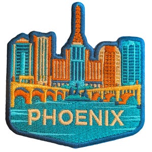 May include: Embroidered patch featuring the Phoenix, Arizona skyline in shades of teal, orange, and yellow. The patch has a shield shape with the city name embroidered in teal at the bottom. The design includes buildings, bridges, and water.