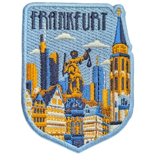 以下が含まれることがあります： 「FRANKFURT」の文字と街並みのイラストが刺繍されたパッチ。青、黄色、オレンジの色合いで建物、像、教会の塔が描かれ、薄い青色の背景になっています。パッチは薄い青色の縁取りがされています。