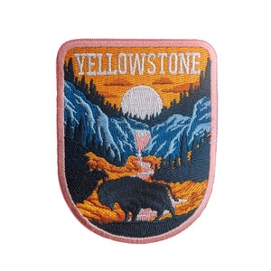 Könnte beinhalten: Ein rosa gestickter Aufnäher mit dem Text "Yellowstone" in Weiß. Der Aufnäher zeigt eine Sonnenuntergangsszene mit einer Bergkette, einem Wasserfall und einer Silhouette eines Bisons.
