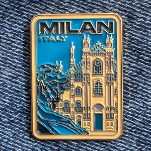Puede incluir: Pin de esmalte dorado con una ilustración azul y blanca de un edificio con el texto "MILAN ITALIA" arriba.