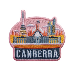 May include: Pink embroidered patch depicting a cityscape of Canberra, Australia. The patch features the Australian Parliament House, a tall tower, and other buildings. The text "CANBERRA" is embroidered in blue on a black background.