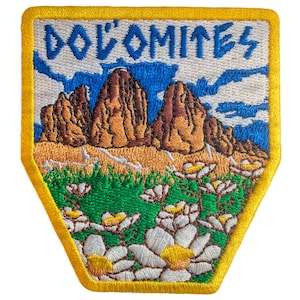 May include: Embroidered patch with the word "DOLOMITES" above a mountain scene. The patch has a yellow border and depicts brown mountains, green grass, white flowers, and a blue sky with clouds. A decorative item.