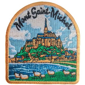 May include: Embroidered patch featuring the Mont Saint Michel in France. The patch has a light blue sky with white clouds, a detailed depiction of the abbey, green grass, blue water, and sheep. The text "Mont Saint Michel" is arched across the top.