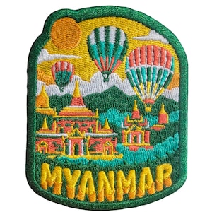 May include: Embroidered patch featuring a scenic Myanmar landscape. The design includes hot air balloons, pagodas, mountains, and the word "MYANMAR" in yellow. The patch has a green border and a yellow and green color scheme.