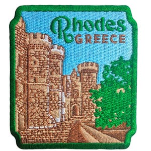 May include: Embroidered patch depicting a castle scene with the text "Rhodes GREECE". The patch has a green border, a blue sky background, and the castle is rendered in shades of brown and tan.