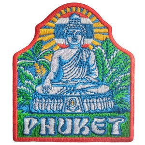 May include: Embroidered patch depicting a seated Buddha in blue and white, set against green foliage and a yellow and blue sky. The word "PHUKET" is embroidered in green at the base, with a red border.
