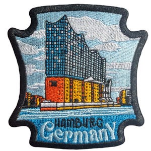 May include: Embroidered patch featuring the Elbphilharmonie concert hall in Hamburg, Germany. The patch has a blue sky background with white clouds, the building is blue and gray, and the base is orange and yellow. The text "Hamburg Germany" is embroidered in blue.