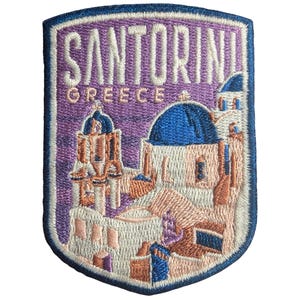 May include: Embroidered patch with a Santorini, Greece design. The shield-shaped patch has a blue border. The design includes the word "SANTORINI" above "GREECE", and depicts the iconic white buildings and blue domes.