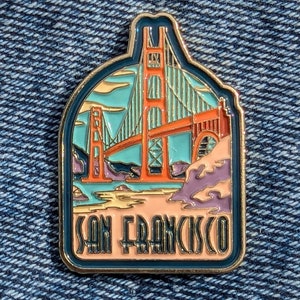 Peut inclure: Une épingle en émail dorée représentant le Golden Gate Bridge de San Francisco en rouge et orange. Le pont est sur un fond bleu et vert avec une plage de couleur pêche au premier plan. Le texte "San Francisco" est écrit en or sous le pont.
