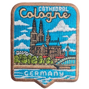 May include: Embroidered patch featuring the Cologne Cathedral in Germany. The patch has a blue sky background with white clouds, the cathedral, a bridge, and the word "Cologne" and "Germany". The patch is framed with a brown border.