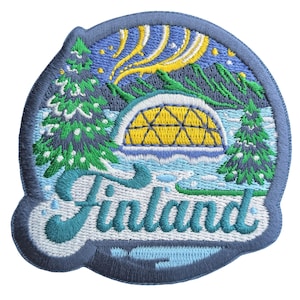 May include: Embroidered patch depicting a Finnish landscape with an igloo, pine trees, and the Aurora Borealis. The word "Finland" is written in a decorative font. The patch has a blue border and is designed to be sewn or ironed onto fabric.