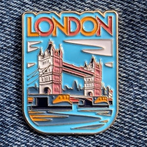 Könnte beinhalten: Eine blaue Emaille-Anstecknadel mit goldener Umrandung, die ein stilisiertes Bild der Tower Bridge in London zeigt. Die Anstecknadel trägt oben den Schriftzug "LONDON" in bunter Schrift.