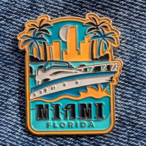 May include: A gold-coloured enamel pin featuring a blue and white boat, palm trees, and the text "MIAMI FLORIDA".