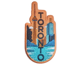 Op de afbeelding: Geborduurde patch met de skyline van Toronto, inclusief de CN Tower en andere gebouwen, met de stadsnaam in oranje hoofdletters. De patch heeft een bruine rand en een blauw-oranje landschap.
