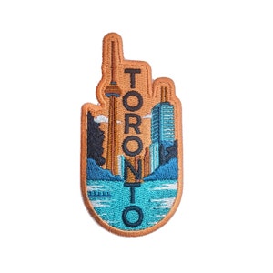 Puede incluir: Parche bordado con el horizonte de Toronto, incluyendo la Torre CN y otros edificios, con el nombre de la ciudad en letras mayúsculas naranjas. El parche tiene un borde marrón y un paisaje azul y naranja.