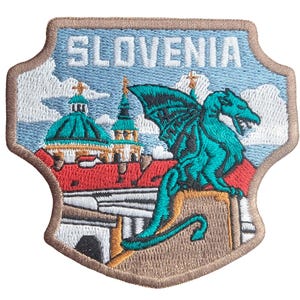 Könnte beinhalten: Gestickter Aufnäher mit dem Wort "SLOVENIA" über einem türkisfarbenen Drachen. Der Aufnäher hat eine Schildform mit hellblauem Hintergrund und einer detaillierten Darstellung eines Gebäudes mit roten Dächern.
