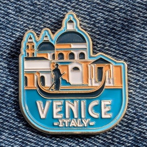 Può includere: Spilla in smalto dorata con un'illustrazione blu e arancione di una gondola a Venezia, Italia. La spilla raffigura un edificio con una facciata bianca e un tetto blu, e il testo "VENICE -ITALY- ".