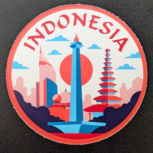 May include: A circular sticker with the word "INDONESIA" in red, encircling a graphic of Indonesian landmarks. The design includes a tall blue monument, a red sun, and other buildings in shades of red, blue, and white.