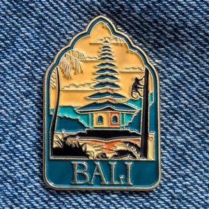 May include: A gold-colored enamel pin with a blue background. The pin depicts a traditional Balinese temple with a yellow roof and green trees. The text "BALI" is written in gold letters at the bottom of the pin.