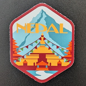 Op de afbeelding: Een zeshoekige sticker met een kleurrijke illustratie van Nepal. Het ontwerp bevat een bergketen, een gouden tempel en het woord "NEPAL" in oranje. De sticker heeft een rode rand en een lichtblauwe achtergrond.