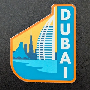 Op de afbeelding: Een kleurrijke sticker met de skyline van Dubai. Het ontwerp omvat het Burj Al Arab hotel, andere gebouwen en het woord "DUBAI" in een verticaal lettertype. De sticker heeft een blauw, geel en oranje kleurenschema.