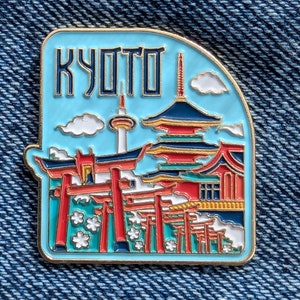 May include: A gold-colored enamel pin with a blue background and the word "KYOTO" in white. The pin depicts a red and gold pagoda with a red torii gate in front of it. There are white clouds and a tower in the background.
