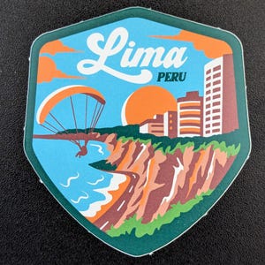 Puede incluir: Una pegatina con forma de escudo que representa una escena costera de Lima, Perú. El diseño incluye un parapente, edificios, una puesta de sol y el texto "Lima PERU" en escritura blanca.