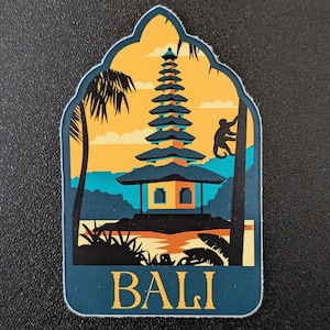 Puede incluir: Una pegatina con una escena de un templo balinés. El diseño incluye una pagoda escalonada, palmeras y una figura trepando a un árbol, sobre un fondo de puesta de sol. La palabra "BALI" está en la parte inferior en color turquesa.