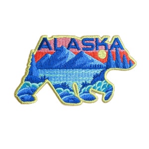 May include: An embroidered patch shaped like a bear with a sunset scene inside. The bear is outlined in yellow thread and the scene includes mountains, a lake, and trees. The word "ALASKA" is stitched in yellow thread across the top of the patch.