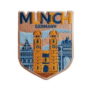 May include: A beige embroidered patch with the text "MUNICH GERMANY" in blue lettering. The patch features a detailed embroidered image of the Frauenkirche church in Munich, Germany.
