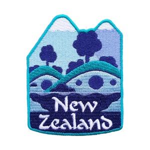 Peut inclure: Un patch brodé avec un motif de paysage bleu et blanc. Le patch représente une chaîne de montagnes, des collines et un lac. Le texte "Nouvelle-Zélande" est brodé en blanc sur un fond bleu.