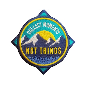 May include: An embroidered patch in blue and yellow with a compass design. The patch features a mountain range with a yellow sun and the text "Collect Moments Not Things".