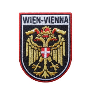 Op de afbeelding: Een geborduurde zwarte en witte patch met een rode rand. De patch heeft een gouden dubbelkoppige adelaar met een rood-wit schild op zijn borst. De tekst "WIEN-VIENNA" staat in wit op een zwarte achtergrond boven de adelaar.