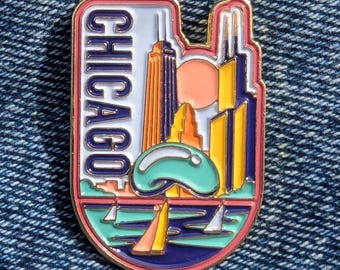 Könnte beinhalten: Ein farbenfroher Emaille-Pin zum Thema Chicago. Der Pin zeigt den Namen der Stadt in weißer, vertikaler Schrift und eine stilisierte Skyline mit Gebäuden, einer Sonne und Segelbooten. Der Pin hat einen rosa Rand und ist vor blauem Denim platziert.