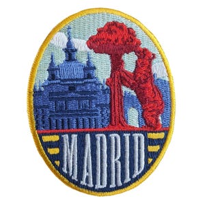 May include: Embroidered patch featuring a blue building, a red bear, and a red tree against a light blue background. The word "MADRID" is displayed in white, with a yellow border. The patch is oval-shaped.