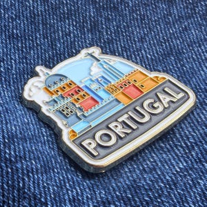 Op de afbeelding: Een rechthoekige pin met een kleurrijke illustratie van Portugese architectuur, met het woord "PORTUGAL" in witte letters op een zwarte achtergrond. De pin heeft een gouden rand en is geplaatst tegen een blauwe denim achtergrond.