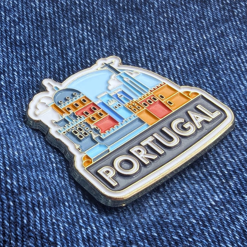 Op de afbeelding: Een rechthoekige pin met een kleurrijke illustratie van Portugese architectuur, met het woord "PORTUGAL" in witte letters op een zwarte achtergrond. De pin heeft een gouden rand en is geplaatst tegen een blauwe denim achtergrond.