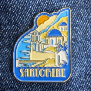 Könnte beinhalten: Eine blaue und goldene Emaille-Anstecknadel mit einer Landschaft von Santorin. Die Nadel zeigt weiße Gebäude mit blauen Kuppeln, eine gelbe Sonne und das Wort "SANTORINI" in blauen Buchstaben. Die Nadel ist vor einem blauen Hintergrund platziert.