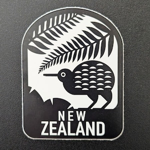 May include: A black and white sticker featuring a kiwi bird, a fern, and the words "NEW ZEALAND". The kiwi bird is depicted with a long beak and a rounded body. The fern is above the bird. The sticker has a rectangular shape with rounded corners.