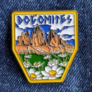 May include: A yellow-edged enamel pin with the word "DOLOMITES" at the top. The pin depicts a mountain range with brown peaks, a blue sky with clouds, and white flowers with yellow centres on a green field.