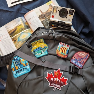 May include: A black backpack with travel patches for Cape Town, Perth, Oslo, Croatia, and Canada. A white Polaroid camera and a travel guide are also visible, suggesting a theme of travel and exploration.