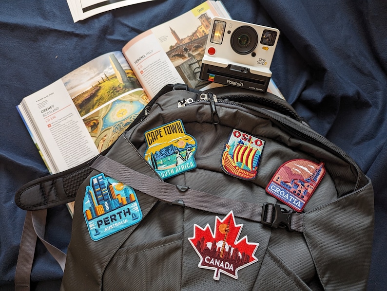 May include: A black backpack with travel patches for Cape Town, Perth, Oslo, Croatia, and Canada. A white Polaroid camera and a travel guide are also visible, suggesting a theme of travel and exploration.