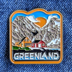 Puede incluir: Pin esmaltado con un paisaje de Groenlandia con montañas, un sol poniente y dos casas. El pin tiene un borde negro con la palabra "GREENLAND" en la parte inferior. El pin está sobre un fondo de mezclilla azul.