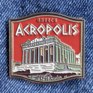 Pode incluir: Um alfinete retangular com a Acrópole de Atenas, Grécia. O alfinete tem um fundo vermelho com a palavra "GREECE" no topo e "ACROPOLIS" em letras brancas. A Acrópole é retratada com colunas cinzentas e verdes e um edifício branco.