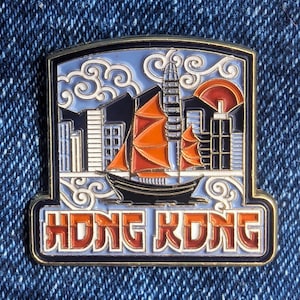 以下が含まれることがあります： 香港の都市景観を描いた長方形のピン。ジャンク船、高層ビル、夕日があります。ピンは金色の縁取りで、「HONG KONG」の文字がオレンジ色で書かれています。