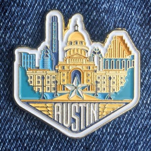 Könnte beinhalten: Ein goldener, weißer und blauer Emaille-Pin mit der Skyline von Austin, Texas. Der Pin enthält den Text "AUSTIN" in einer stilisierten Schriftart, einen Stern und ikonische Gebäude. Der Pin ist vor einem Jeans-Hintergrund platziert.