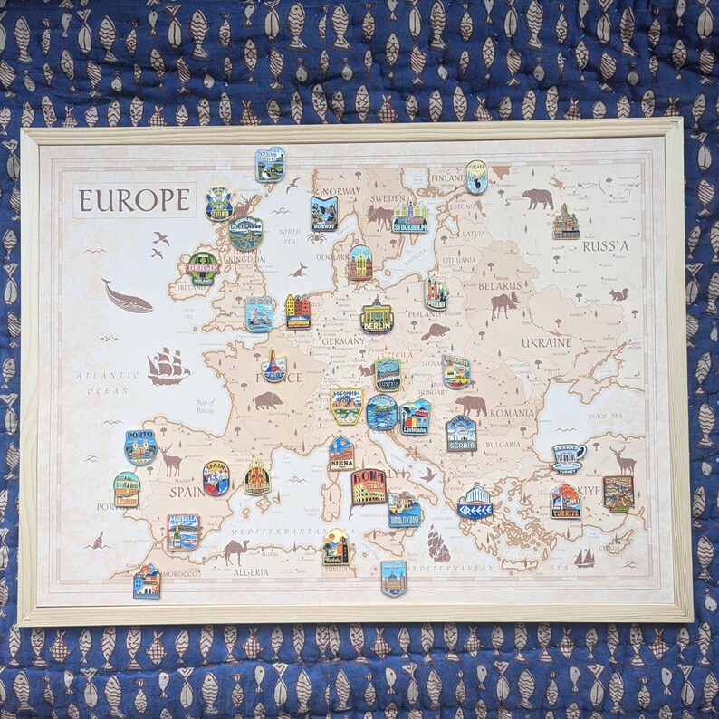 K&ouml;nnte beinhalten: Eine gerahmte Europakarte mit Pins, die verschiedene Orte markieren. Die Karte ist beige mit dem Wort "EUROPE" oben. Pins mit farbenfrohen Designs sind auf verschiedenen L&auml;ndern platziert. Der Hintergrund ist ein blauer Stoff mit Fischmuster.