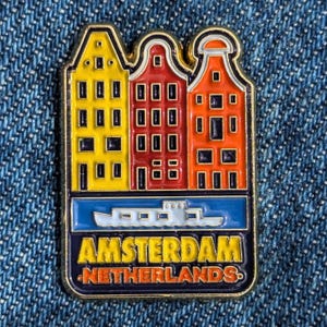 Puede incluir: Un pin rectangular de esmalte con tres casas de canal coloridas en amarillo, rojo y naranja. Debajo, una sección azul representa un barco, con las palabras "AMSTERDAM NETHERLANDS" en amarillo.